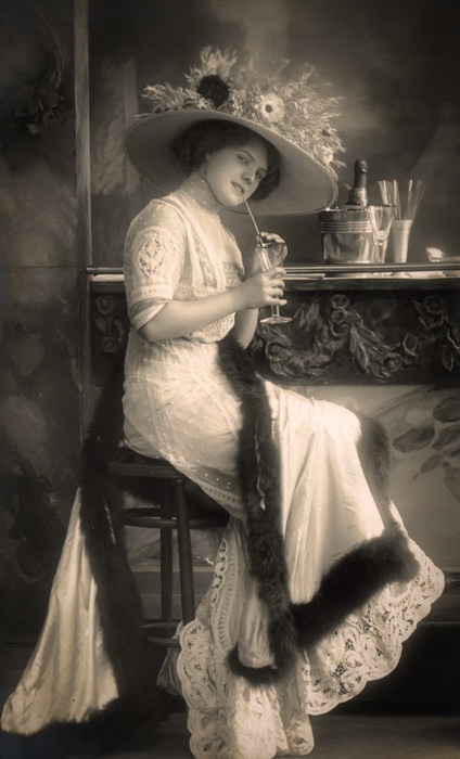 Élise – La robe à franges Élise est vêtue d'une élégante robe de dentelle et est coiffée d'un large chapeau fleuri. Cette photographie ancienne met en valeur le raffinement de la mode féminine au début du XXᵉ siècle.