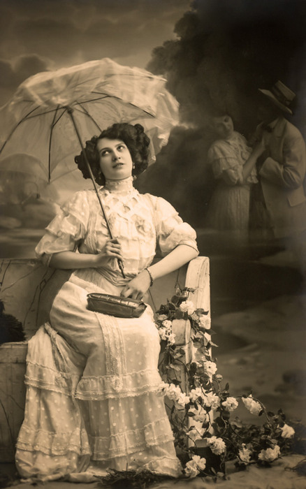 Germaine songeant – L'amour imaginé vers 1900 Dans cette carte postale de la Belle Époque, une jeune femme songe sous son ombrelle, tandis qu’un couple amoureux apparaît en arrière-plan. Le jeu de transparence transforme cette scène en une évocation délicate du rêve amoureux.