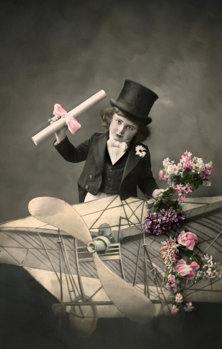 Arthur – Un message pour vous ! Arthur brandit un petit rouleau de papier à bord d’un aéroplane factice décoré de fleurs. Cette mise en scène photographique témoigne de la fascination pour l’aviation et des rêves de voyage qui nourrissaient l’imaginaire du début du XXᵉ siècle.