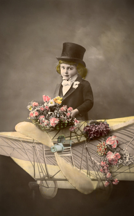 Arthur – Cap sur les nuages Arthur pose dans un aéroplane reconstitué en studio, entouré de bouquets de fleurs. Cette carte postale ancienne témoigne de la fascination pour l’aviation et du rêve de voyage qui accompagne les débuts de l’aéronautique.
