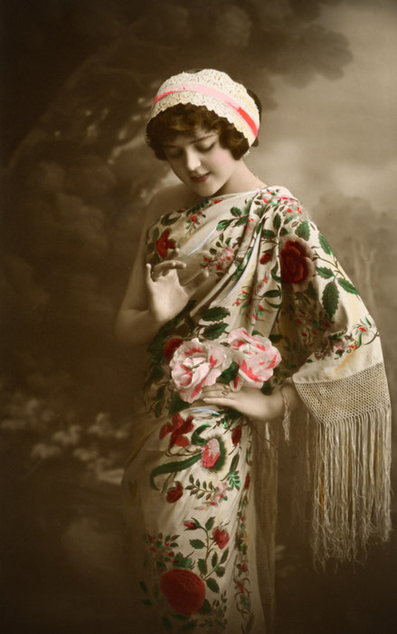 Eva – Le châle brodé de fleurs Eva porte un grand châle à franges richement brodé de motifs floraux. Elle porte également un bonnet de dentelle. Cette photographie ancienne évoque le raffinement de la mode féminine des années 1910.