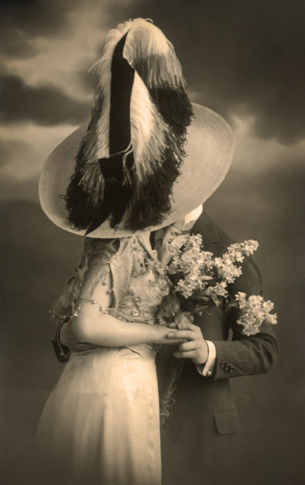 Louise et Joseph – Derrière le grand chapeau Louise et Joseph se cachent derrière un immense chapeau orné de plumes pour s'embrasser. Cette carte postale photographique ancienne évoque les amours discrètes de la Belle Époque.