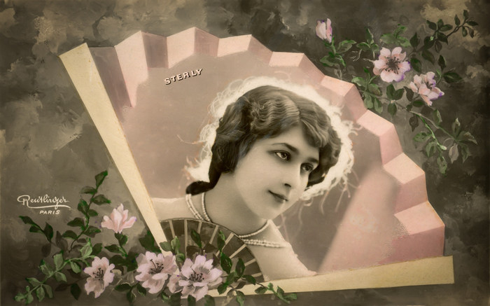 L'éventail – Sterly vers 1900 Dans cette carte postale de la Belle Époque, Sterly apparaît au cœur d’un éventail délicatement orné de fleurs. Les plis de l’éventail encadrent son portrait tandis que les motifs floraux prolongent cette composition raffinée.