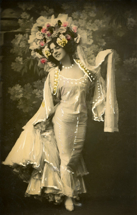 Olga et sa couronne de fleurs Olga porte une robe fluide aux manches longues, de même qu'une spectaculaire couronne de fleurs qui encadre son visage. Cette carte postale ancienne célèbre l'élégance des costumes de scène de la Belle Époque.