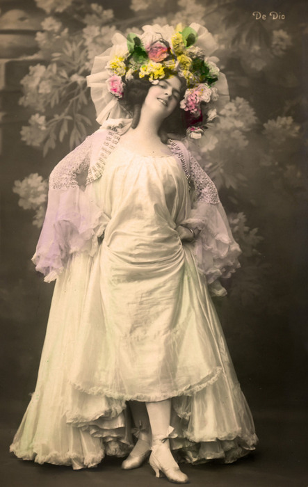 Olga – Les voiles pastel Olga pose vêtue d'une robe claire agrémentée de voiles transparents. Elle est coiffée d'une imposante couronne de fleurs. Cette photographie de la Belle Époque met en valeur la mode féminine des années 1900.