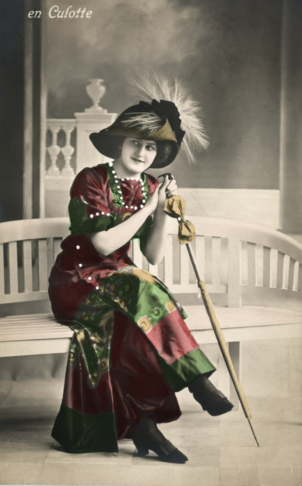 La jupe-culotte de Renée Assise sur un banc, Renée porte une jupe-culotte, un haut assorti et un grand chapeau orné de plumes. Cette carte postale illustre une mode féminine novatrice au début du XXᵉ siècle.