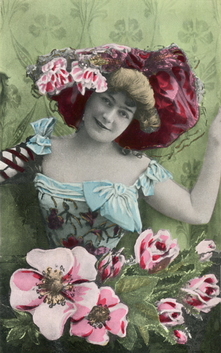 Félicie s'épanouit parmi les fleurs Dans cette composition photographique colorisée de la Belle Époque, Félicie apparaît entourée de fleurs et coiffée d’un imposant chapeau orné de roses. Les fleurs du premier plan et les détails colorés de sa tenue composent un ensemble particulièrement foisonnant.