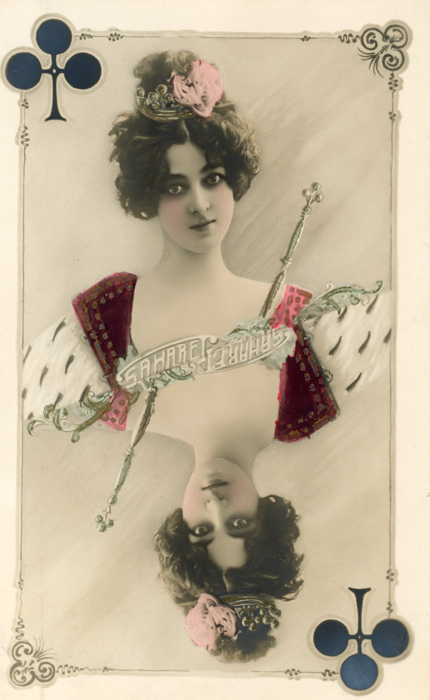 Saharet incarne la dame de trèfle Dans cette carte postale photographique de la Belle Époque inspirée des cartes à jouer, la danseuse Saharet apparaît sous les traits de la dame de trèfle. La composition associe son portrait aux symboles et aux ornements caractéristiques du jeu de cartes.
